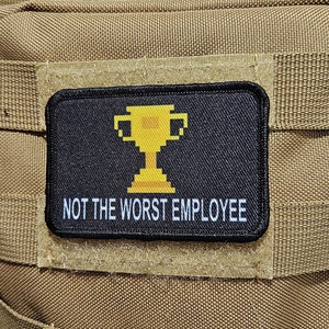 May include: A tan tactical backpack with two patches. The top patch is a black and white American flag. The bottom patch is black with a yellow pixelated trophy and the text "NOT THE WORST EMPLOYEE".