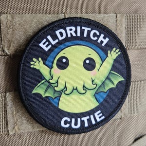 May include: A circular patch with the text "ELDRITCH CUTIE" in white. The patch depicts a cartoon green creature with wings and tentacles, set against a blue and black backdrop. An American flag patch is visible above.