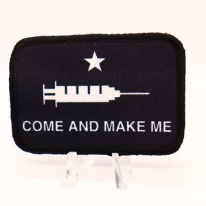 May include: A black and white patch with a white star and a syringe with the text "COME AND MAKE ME" on a black background.