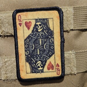 May include: A rectangular patch featuring a playing card design, the Queen of Hearts, with a skeletal figure wearing a crown. The card has a tan background with black and red details. The patch is bordered in black and is attached to a tan fabric.