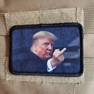 May include: A rectangular patch featuring a portrait of a man in a suit, giving the middle finger. The patch is on a tan fabric background, with a black and white American flag patch above it. The patch is likely an accessory.