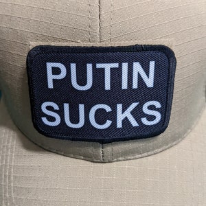 May include: A tan baseball cap with a black patch that says "PUTIN SUCKS" in white letters.