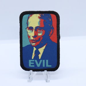 Op de afbeelding: Een geborduurde zwarte en witte patch met een rode, blauwe en gele afbeelding van een man met een bril. De tekst "EVIL" is in groen onder de afbeelding gedrukt.