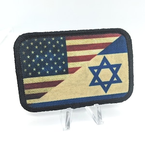 Può includere: Una toppa con bordo nero che mostra una bandiera americana sbiadita e una bandiera israeliana. Le bandiere sono cucite insieme con la bandiera americana in alto e la bandiera israeliana in basso.