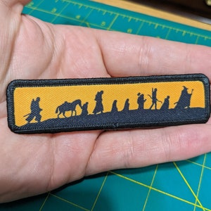 Puede incluir: Un parche bordado negro y amarillo con la silueta de un grupo de figuras caminando por un sendero. Las figuras caminan en fila y el sendero está delineado en negro. El parche es rectangular y tiene un borde negro.