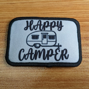 Könnte beinhalten: Ein weißer Patch mit schwarzer Stickerei, auf dem "Happy Camper" steht, mit einer kleinen Camper-Illustration in der Mitte.