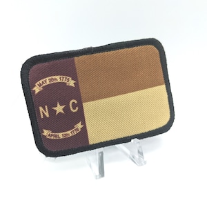 Op de afbeelding: Een bruine, witte en bordeauxrode stoffen patch met de vlag van de staat North Carolina. De vlag toont een witte ster op een bordeauxrode achtergrond met de letters "N.C." aan weerszijden van de ster. De vlag heeft ook een banner met de tekst "MAY 20th 1775" en "APRIL 12th 1776".