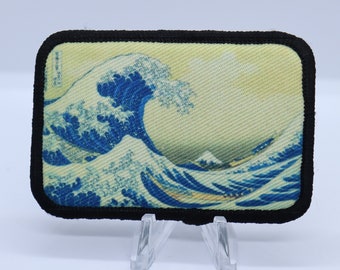 May include: A black framed patch with a printed image of the Great Wave off Kanagawa by Katsushika Hokusai. The wave is blue and white with a yellow background.