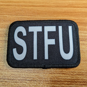 May include: A black patch with white letters that spell out "STFU".