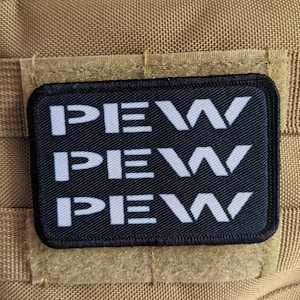 Könnte beinhalten: Ein schwarzer Patch mit weißem Text, der "PEW PEW PEW" lautet, ist an einer braunen Stoffoberfläche befestigt.
