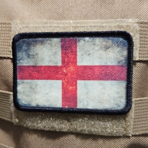 May include: A white and red patch with a cross design, sewn onto a brown tactical vest. The patch has a distressed, vintage look.