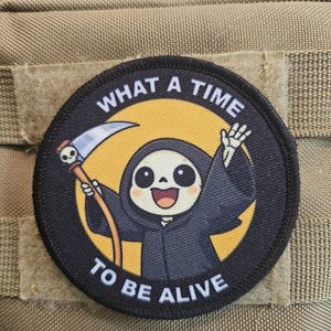 Può includere: Toppa rotonda nera con la scritta "WHAT A TIME TO BE ALIVE" che circonda un personaggio dei cartoni animati di Grim Reaper. Il Reaper sorride e tiene una falce. La toppa è su un tessuto beige.
