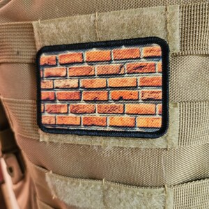 May include: A brown brick pattern patch with black stitching around the edges. The patch is attached to a tan tactical vest.