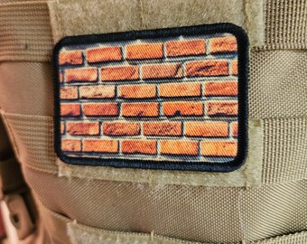 May include: A brown brick pattern patch with black stitching around the edges. The patch is attached to a tan tactical vest.