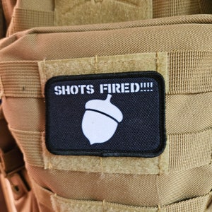 May include: A black patch with white text that reads "SHOTS FIRED!!!" and a white acorn graphic.