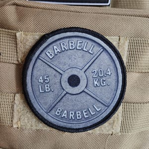 May include: A round patch featuring a barbell weight plate design. The patch is gray with black text that reads "BARBELL", "45 LB.", and "20.4 KG.". The patch is attached to a tan fabric.