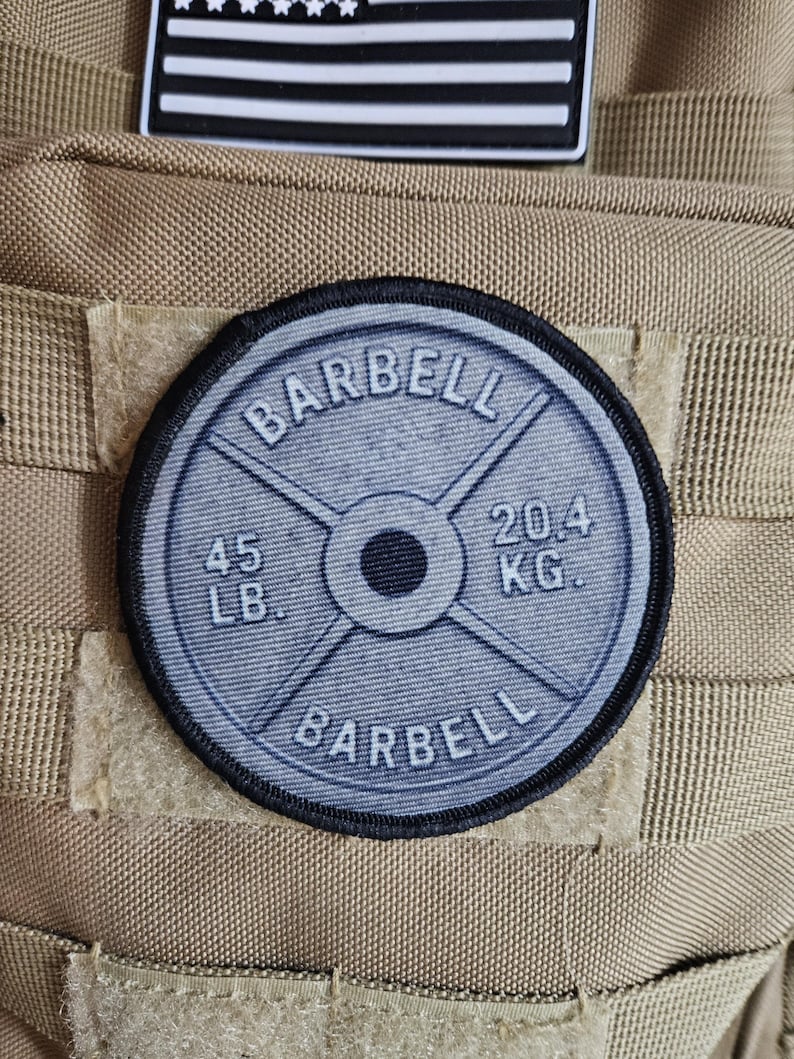 May include: A round patch featuring a barbell weight plate design. The patch is gray with black text that reads "BARBELL", "45 LB.", and "20.4 KG.". The patch is attached to a tan fabric.