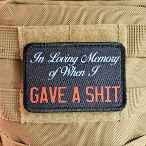 May include: A black rectangular patch with the text "In Loving Memory of When I GAVE A SHIT" in white and orange lettering. The patch is attached to a tan fabric background, with a black and white striped patch above it.