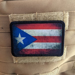 Pode incluir: Um remendo da bandeira de Porto Rico desgastado, desbotado e desfiado com uma borda preta. O remendo está preso a um tecido bege texturizado com velcro.