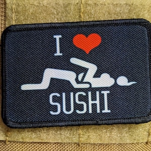 Könnte beinhalten: Ein schwarzer Patch mit weißem Text, der "Ich liebe Sushi" sagt und ein rotes Herz. Der Text ist in einer stilisierten Schrift. Der Patch hat eine weiße Silhouette einer Person in einer suggestiven Pose.
