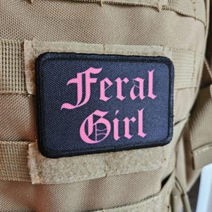 Könnte beinhalten: Ein schwarzer Patch mit pinkfarbener Aufschrift "Feral Girl" in gotischer Schrift.