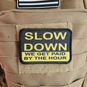 Può includere: Uno zaino tattico color beige con una toppa della bandiera americana in bianco e nero e una toppa gialla e nera che recita "Slow Down We Get Paid By The Hour."