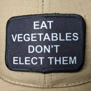 May include: A black patch with white text that reads "EAT VEGETABLES DON'T ELECT THEM" on a tan baseball cap.