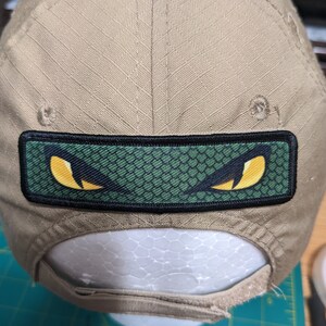 Puede incluir: Una gorra de béisbol de color beige con un parche de ojo de serpiente negro y verde. El parche tiene un patrón de piel de serpiente verde con dos ojos amarillos.