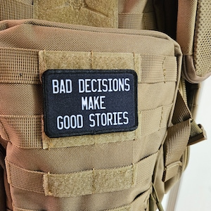 May include: A black patch with white text that reads "Bad Decisions Make Good Stories" is sewn onto a tan tactical backpack.