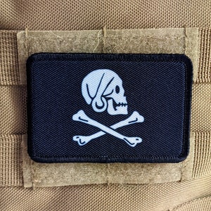 May include: A black patch with a white skull and crossbones. The skull is wearing a white bandana. The patch is sewn onto a tan fabric background.