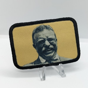 May include: A black and yellow embroidered patch featuring a portrait of a man wearing glasses and a smile. The patch is on a white background.