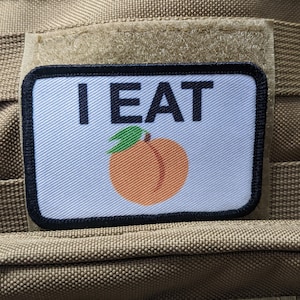 Puede incluir: Un parche blanco con costuras negras y texto negro que dice "I EAT" con una ilustración de melocotón. El parche está unido a un fondo de tela marrón.