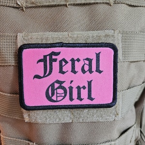 May include: A pink patch with black embroidered text that reads "Feral Girl" on a tan background.