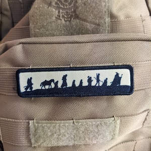 Puede incluir: Un parche blanco con costuras negras presenta una silueta de un grupo de figuras caminando por un bosque. Las figuras incluyen una persona montando un caballo, una persona llevando un bastón y una persona con un sombrero. El parche está unido a un fondo de tela marrón.