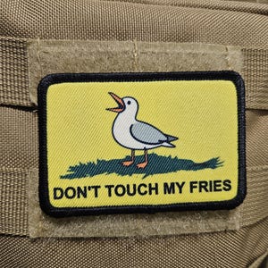 May include: A rectangular patch with a yellow background and a black border. The patch features a cartoon seagull with its beak open, standing on green grass. The text "DON'T TOUCH MY FRIES" is below the seagull.