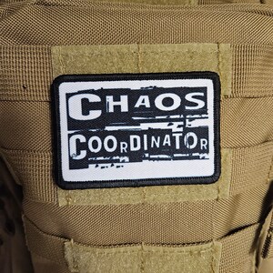 Peut inclure: Un patch blanc avec du texte noir qui dit "CHAOS COORDINATOR" sur un fond brun.