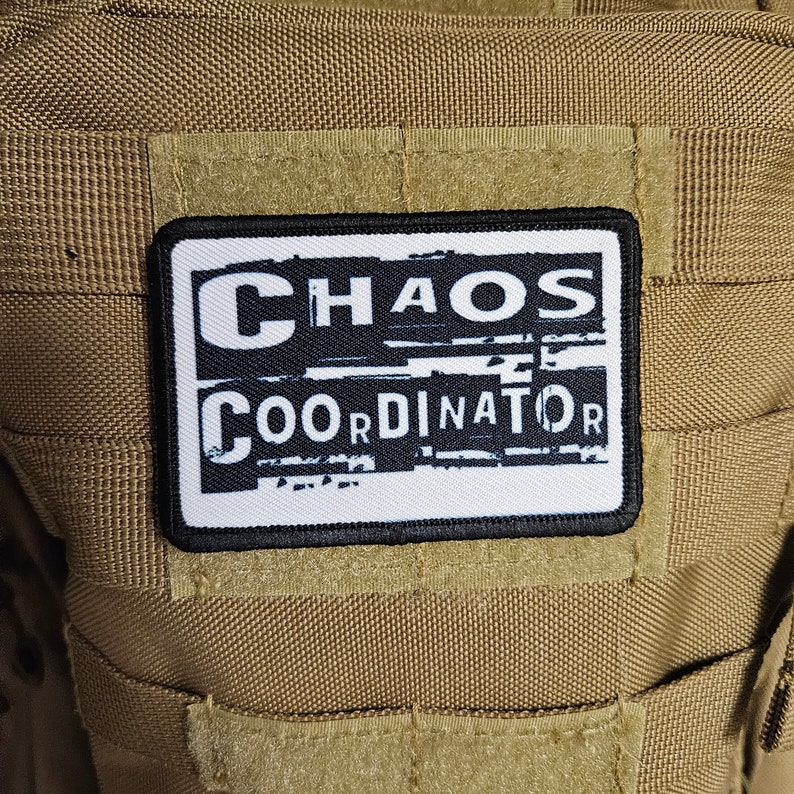 Peut inclure: Un patch blanc avec du texte noir qui dit "CHAOS COORDINATOR" sur un fond brun.