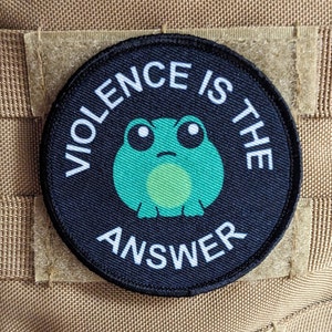 Könnte beinhalten: Ein schwarzer und weißer runder Patch mit einem grünen Frosch in der Mitte. Der Text "Violence is the answer" ist um den Frosch herum geschrieben.