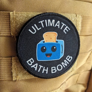 Könnte beinhalten: Ein runder, schwarzer Aufnäher mit den weißen Worten "ULTIMATE BATH BOMB". Der Aufnäher zeigt einen blauen Toaster mit einer Toastscheibe und einem lächelnden Gesicht. Der Aufnäher ist an einem beigefarbenen Stoff befestigt.