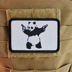 May include: A black and white panda bear holding two guns, printed on a white patch with a black border. The patch is attached to a brown tactical vest.