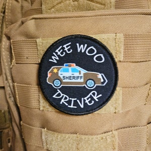 Puede incluir: Un parche blanco y negro con las palabras "Wee Woo Driver" y una imagen de dibujos animados de un coche de sheriff con una estrella en el lateral y la palabra "Sheriff" en la puerta.