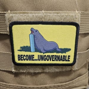May include: A rectangular morale patch with a yellow background and a black border. The patch features a purple seal biting a light blue rectangular object, set against a green patch of grass. The text BECOME...UNGOVERNABLE is printed in black.