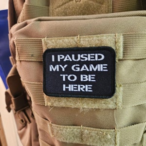 May include: A black patch with white text that reads "I paused my game to be here" is attached to a tan tactical backpack.
