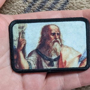 May include: A black and white embroidered patch featuring a portrait of a man with a long white beard and a white robe. The man is holding a staff in his right hand.