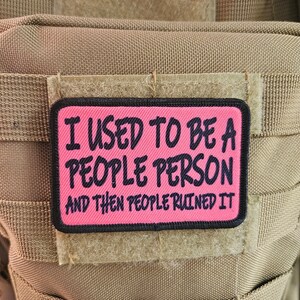 May include: A rectangular patch with a pink background and black border. The patch reads "I USED TO BE A PEOPLE PERSON AND THEN PEOPLE RUINED IT" in black text. The patch is attached to a tan fabric.