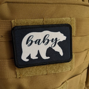 May include: A black and white patch with a white bear silhouette and the word "baby" written in cursive. The patch is attached to a brown fabric background.