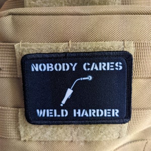 Può includere: Toppa bianca e nera con il testo "Nobody Cares Weld Harder" e una silhouette bianca di una torcia da saldatura.