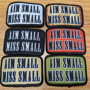 Peut inclure: Six patchs brodés avec le texte "Aim Small Miss Small" en lettres noires sur des fonds de couleurs différentes. Les patchs sont blancs, orange, noirs, rouges, beiges et verts.