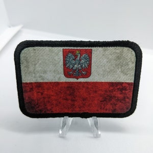 May include: A distressed, embroidered patch featuring the Polish flag. The flag is red and white with the Polish coat of arms in the upper left corner. The patch is surrounded by a black border.