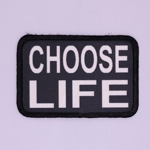 Op de afbeelding: Zwarte stoffen patch met witte tekst die "CHOOSE LIFE" luidt.